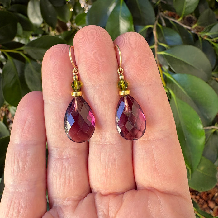 A pair of teardrop-shaped dangling earrings with faceted burgundy-purple stones and small gold-toned beads, held between a person's fingers with green leaves in the background.
