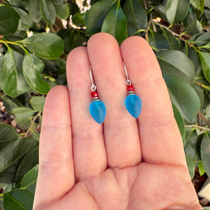 A pair of dangling earrings with light blue teardrop-shaped beads and small red beads, held between fingers against a background of green leaves.