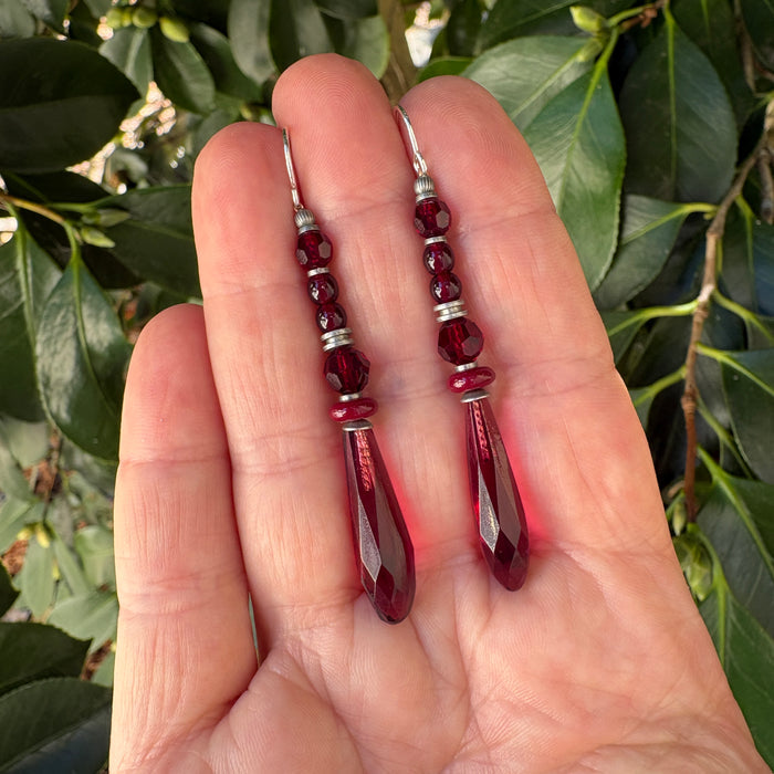 A pair of dangling earrings with faceted dark red beads and silver-toned spacers, held in a person's hand against a background of green leaves.