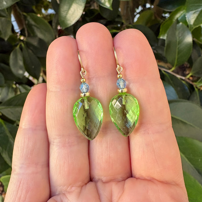A pair of gold drop earrings with faceted light green teardrop-shaped gemstones and small round blue beads, held between a person's fingers with green leaves in the background.