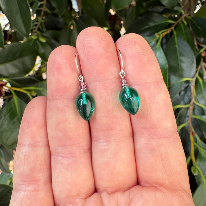 A pair of green teardrop-shaped glass drop earrings with silver fishhook backs, held on a person's fingers against a background of green leaves.