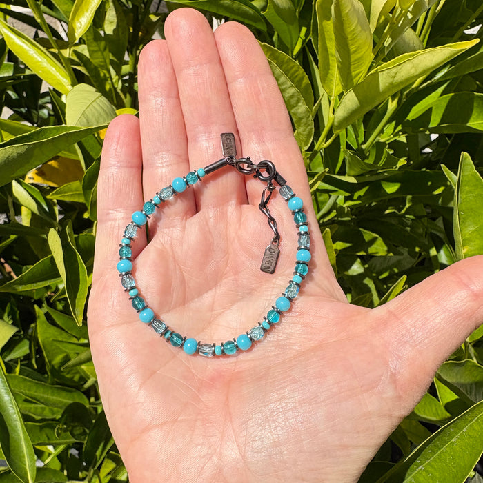 A beaded bracelet with turquoise blue and clear faceted beads on a dark metal clasp, held in a person's hand with green leaves in the background.