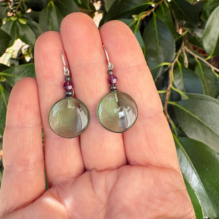 A pair of handmade dangle earrings held in a person's hand, featuring round olive-green cabochon stones and small purple beads, set against a background of green leaves.